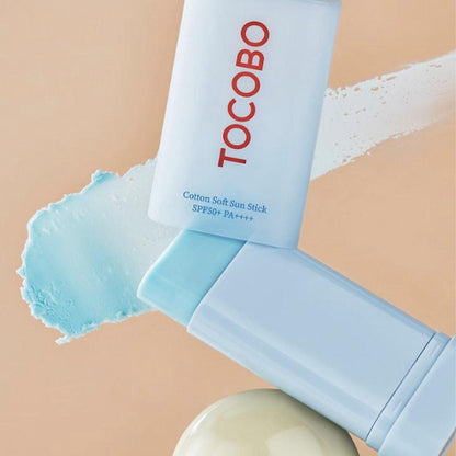 Two light blue Tocobo Cotton Soft Sun Sticks (SPF50+ PA++++), featuring red TOCOBO text, are set against a beige background. A matching blue smear extends from the top right, with a beige round object below, emphasizing this 19g vegan sun sticks charm.