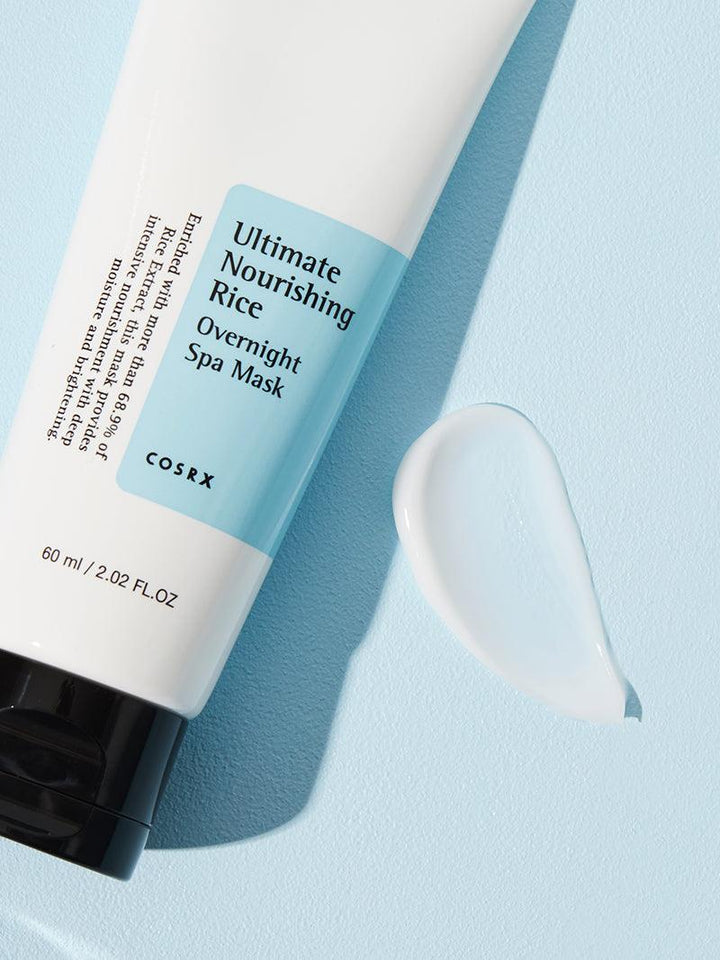 A white tube of COSRX Ultimate Nourishing Rice Overnight Spa Mask - 60ml featuring a black cap and light blue label lies on a light blue surface, with a dollop of the brightening rice extract-infused moisturizer beside it.