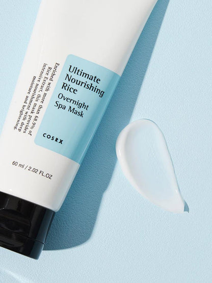 A white tube of COSRX Ultimate Nourishing Rice Overnight Spa Mask - 60ml featuring a black cap and light blue label lies on a light blue surface, with a dollop of the brightening rice extract-infused moisturizer beside it.