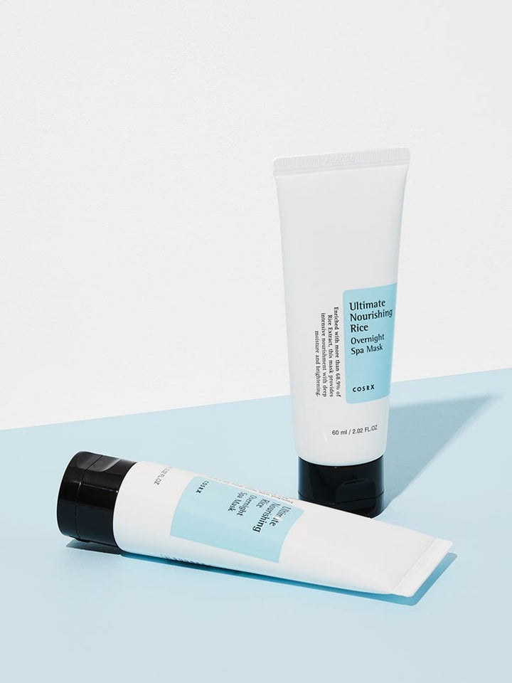 Two white tubes with black caps are set against a light blue and white background. One stands upright while the other lies down. Labeled COSRX Ultimate Nourishing Rice Overnight Spa Mask - 60ml, it features rice extract for moisturizing and brightening effects.