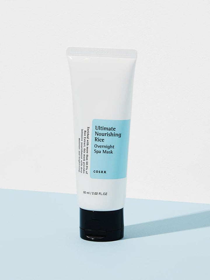 A white tube with a black cap stands upright on a two-toned blue and white background. The label reads COSRX Ultimate Nourishing Rice Overnight Spa Mask - 60ml, featuring rice extract for brightening and deep moisturizing. Net weight: 60 ml / 2.02 FL.OZ.